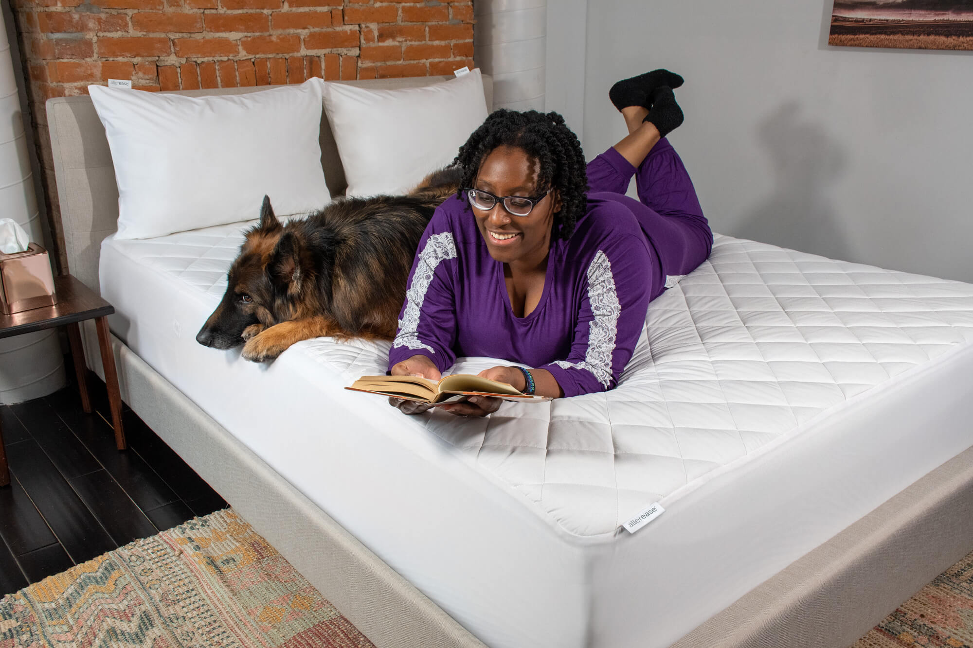 A woman reading a book with her german shepard laying on a mattress pad.
