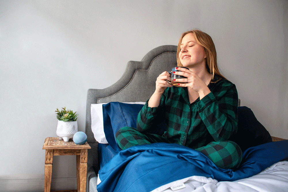 Woman sitting on allerease bedding drinking tea and closing eyes